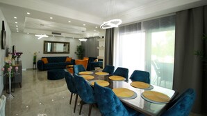 A modern living room with a dining area, featuring a glass-top table, blue upholstered chairs, and a large window with curtains.
