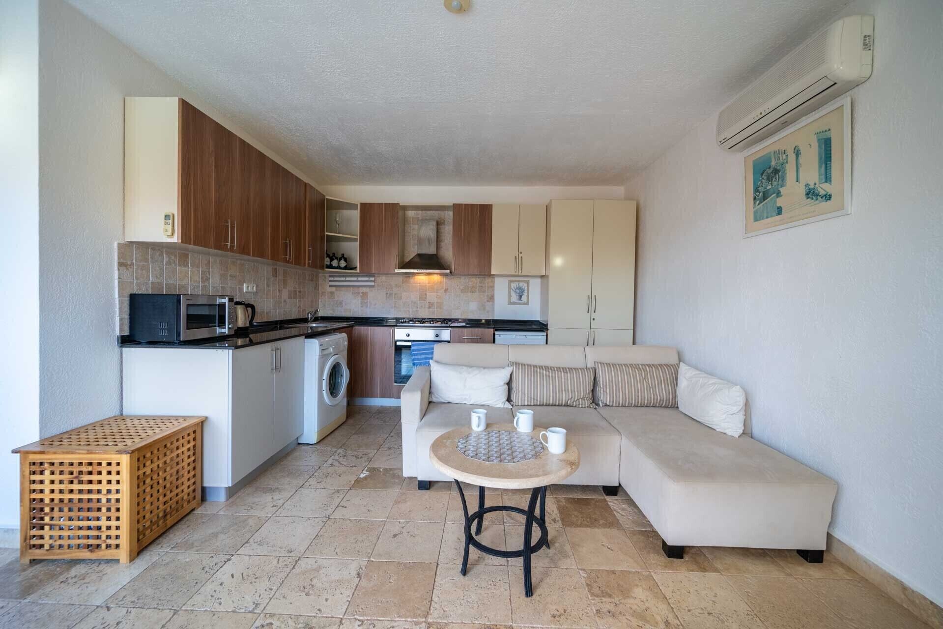 A compact kitchen with a washing machine, a microwave, and a stove. There is a sofa and a small round table with cups.