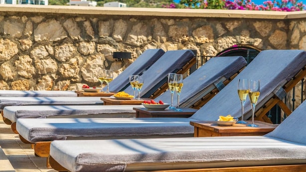 Une rangée de chaises longues, avec des serviettes et des verres de vin blanc, est disposée devant un mur de pierre, offrant une vue sur une colline et des fleurs.