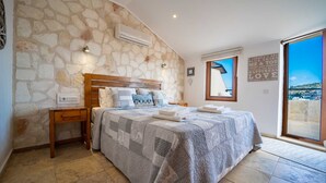 Une chambre avec un mur en pierre, une tête de lit en bois, un lit recouvert d’une couvre-lit à motifs, une table de chevet, une fenêtre avec vue et un mur orné d’une citation inspirante.