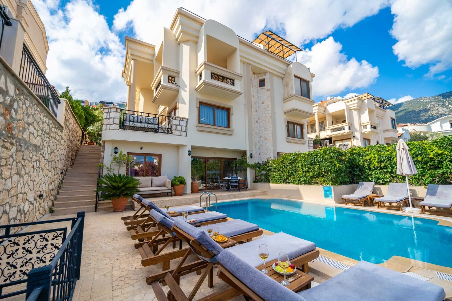 A poolside area with lounge chairs and a swimming pool, surrounded by a multi-story building with balconies and a stone wall.