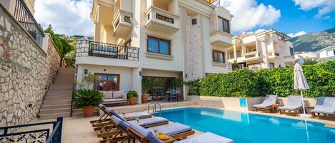 A poolside area with lounge chairs and a swimming pool, surrounded by a multi-story building with balconies and a stone wall.