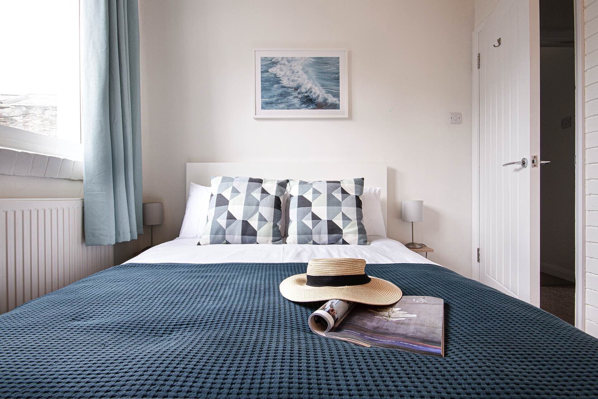 A neatly made bed with a patterned pillow, a hat, and a magazine on a textured bedspread. A framed picture hangs on the wall above the bed.