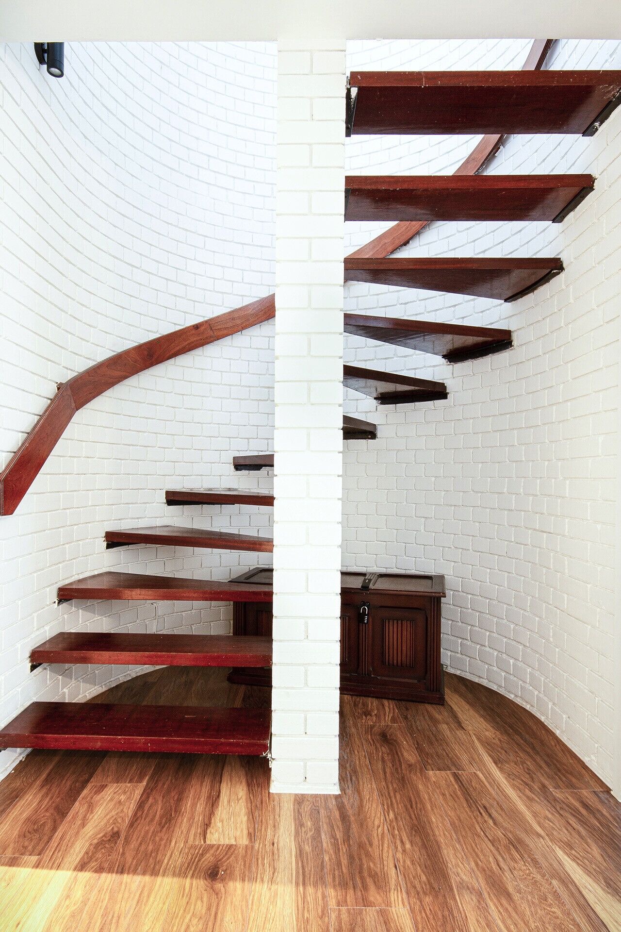 A spiral staircase with wooden steps and beams against a white brick wall, leading to a dark wooden cabinet.