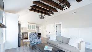 Un vaste salon comprenant un canapé, une table basse et un fauteuil à motifs. On aperçoit, au fond, une cuisine avec des meubles blancs et une cuisinière.