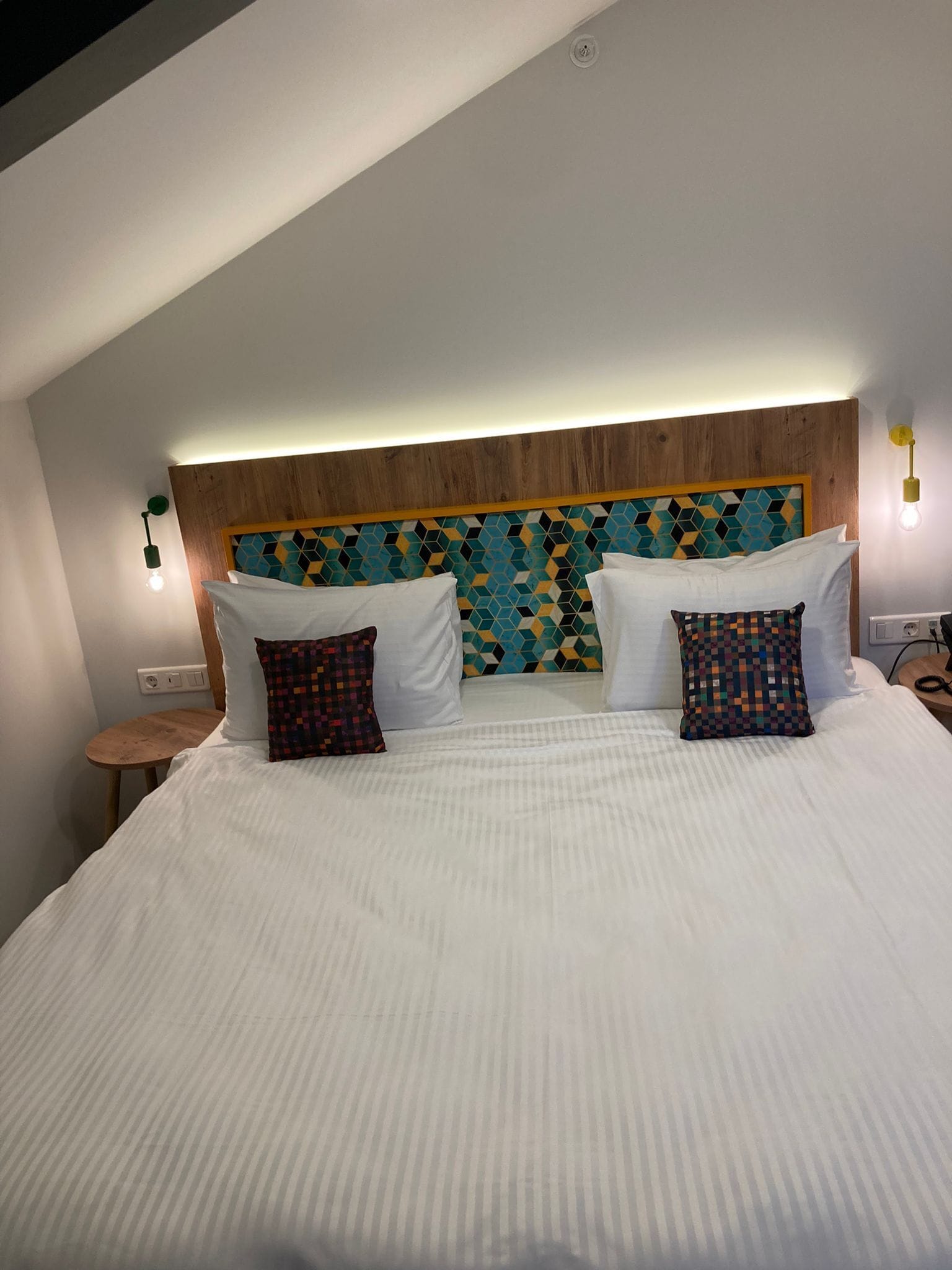 A neatly made bed with white linens and two patterned pillows, a wooden headboard with a geometric tile design, and a small wooden side table.