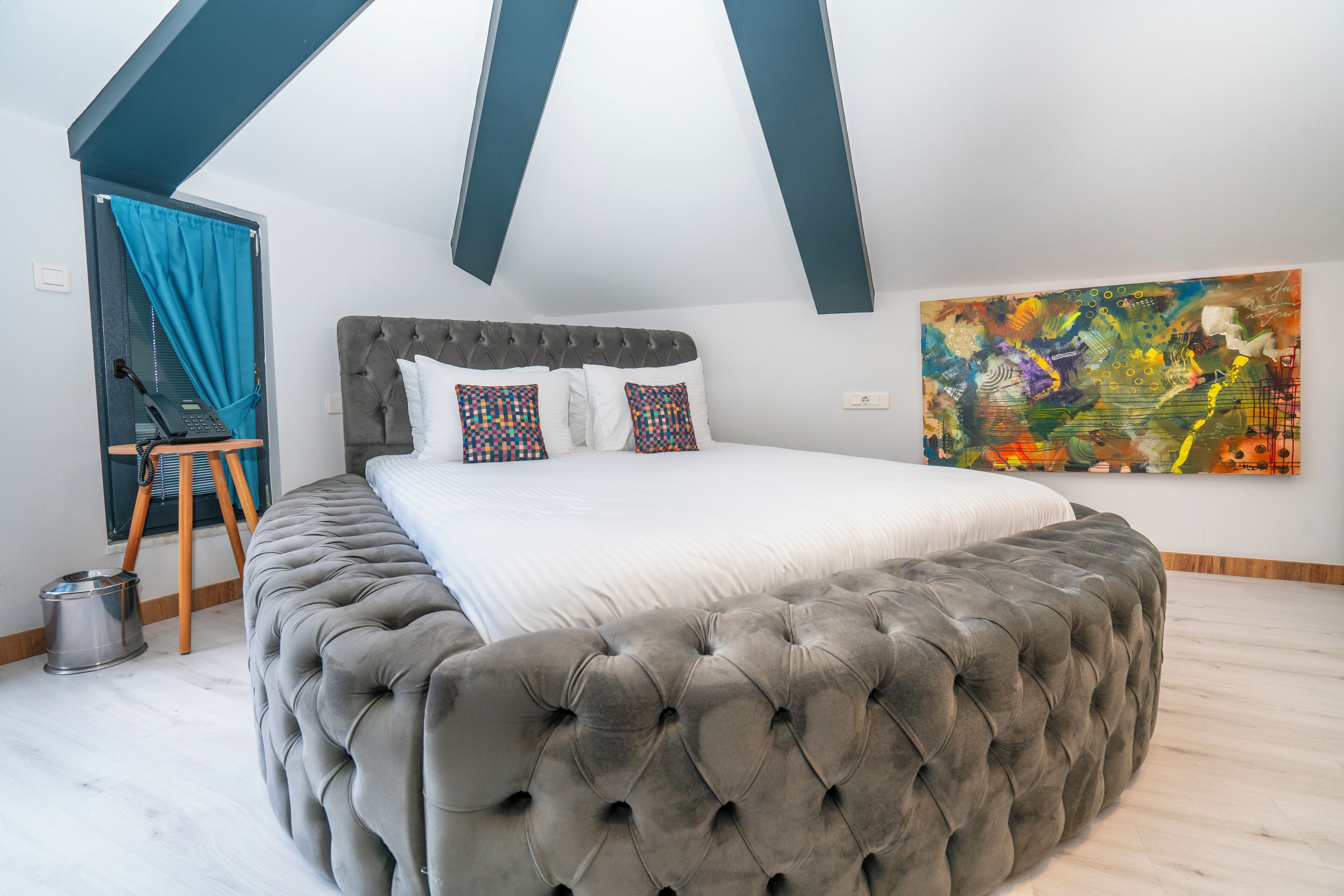 Ein Schlafzimmer mit einer gesteppten Kopfteil, einem großen Bett, einem farbenfrohen abstrakten Gemälde und einem Fenster mit blauer Vorhang.