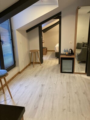 Ein modernes, gut beleuchtetes Zimmer mit Holzboden, einem kleinen Hocker, einem Bett mit weißer Bettdecke, einem Minikühlschrank und einer Sitzecke mit einer Couch.