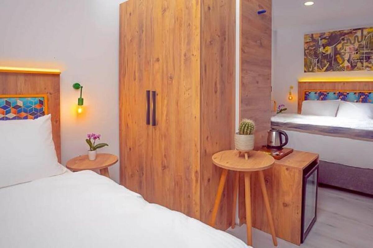 Habitación de hotel moderna con armario de madera, cama con cabecera colorida, una pequeña mesa con una planta y una obra de arte colgada en la pared.