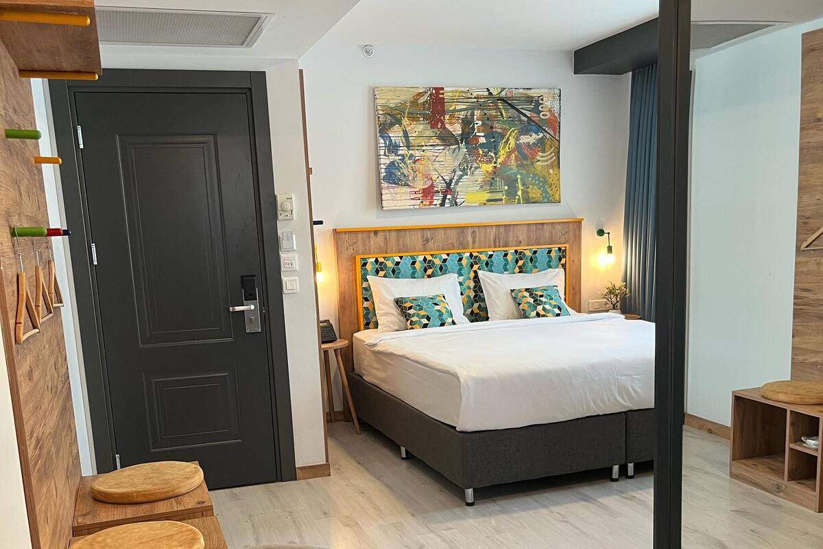 Una habitación de hotel moderna con una cama, una silla, una mesita con una planta, una puerta y un cuadro en la pared.