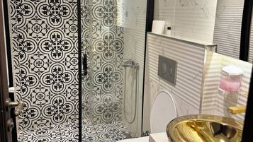 Un baño con piso estampado, un inodoro, un lavamanos con lavabo dorado y una zona de ducha con puerta de cristal.