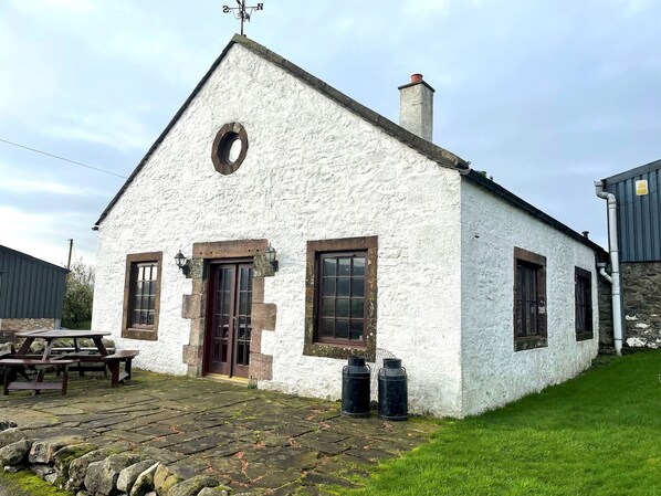 A white building with a pitched roof, a chimney, and a circular window. There are two doors and two windows with shutters. A picnic table is outside the building.