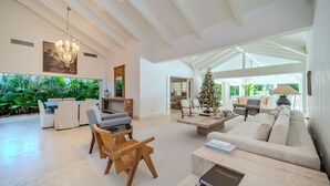 Un vaste espace de vie comprenant un grand canapé, une table basse et un coin repas avec des chaises. Un lustre est suspendu au plafond et un sapin de Noël se trouve au fond de la pièce.