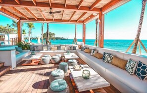 A covered outdoor seating area with a view of the ocean, wooden flooring, and a variety of seating options including sofas and round tables. - Chic Villa in Casa de Campo_3 mins from the beach ! (La Romana)