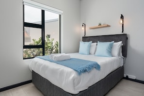 A neatly made bed with a grey headboard, white bedding, and a blue throw blanket. There is a window with a view of greenery outside. - Light and Cozy Blouberg Apartment (Cape Town)