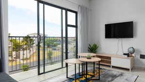 A modern living room with a large sliding glass door to a balcony, a flat-screen TV mounted on the wall, and a wooden console table with a small plant. - Light and Cozy Blouberg Apartment (Cape Town)