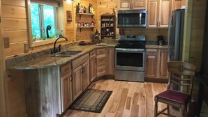 A wooden cabin kitchen with a granite countertop, stainless steel appliances, and a wooden chair. - Riverfront! Private, short walk to town (Hot Springs)
