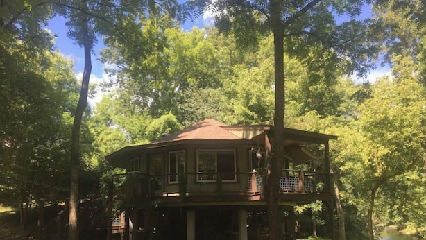 A two-story wooden cabin surrounded by trees, with a balcony on the upper floor. - Riverfront! Private, short walk to town (Hot Springs)