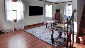 A living room with hardwood flooring, a large flat-screen TV mounted on the wall, a wooden coffee table, and a staircase with a wooden railing. - The Arch House (Faribault)