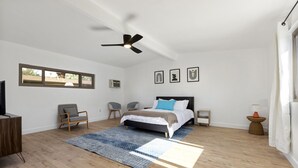 A modern bedroom with a bed, two chairs, a TV, a ceiling fan, and a window with a view. - Private mini oasis steps from El Paseo! (Palm Desert)