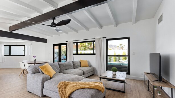 A modern living room with a sectional sofa, a coffee table, a TV, and large windows with curtains. - Private mini oasis steps from El Paseo! (Palm Desert)