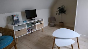 A modern living room with a white TV stand, a small table, a wicker chair, and a potted plant. - "Le Gite tranquille" by the sea in Surtainville (Surtainville)
