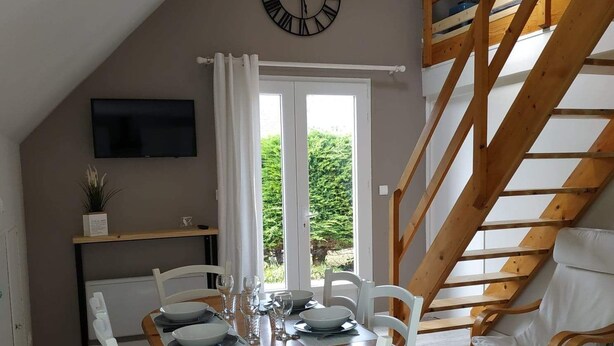A dining area with a wooden table set for four, white chairs, a clock on the wall, and a staircase leading to an upper level. - "Le Gite tranquille" by the sea in Surtainville (Surtainville)