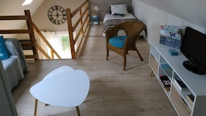 A cozy attic bedroom with a sloped ceiling, a wooden chair, a white table, a bed with a blue cushion, and a TV on a white shelf. - "Le Gite tranquille" by the sea in Surtainville (Surtainville)