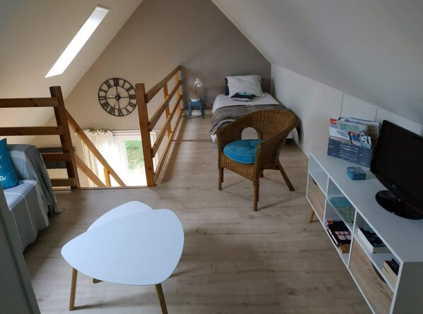 Un dormitorio acogedor en el ático, con techo inclinado, una silla de madera, una mesa blanca, una cama con cojín azul y un televisor sobre un estante blanco.