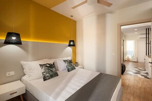 A modern bedroom with a bed, bedside tables, and a nightstand. There is a hallway leading to another room. - Salty Colours Apartments in Almyrida (000)