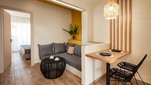 A modern living room with a sofa, a round black wicker coffee table, a wooden dining table with two chairs, and a hanging light fixture. - Salty Colours Apartments in Almyrida (000)