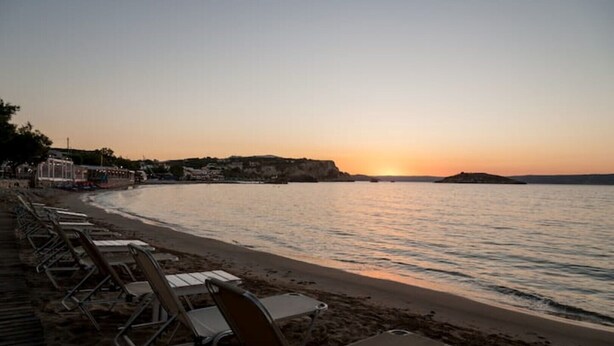 A beach with rows of empty sun loungers facing the sea, a clear sky, and a distant sunset. - Salty Colours Apartments in Almyrida (000)