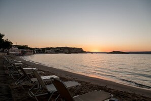 A beach with rows of empty sun loungers facing the sea, a clear sky, and a distant sunset. - Salty Colours Apartments in Almyrida (000)