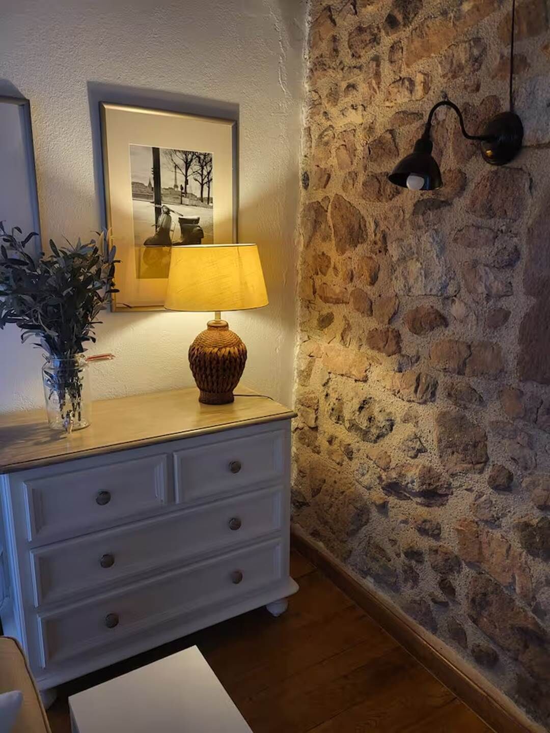 Une pièce avec un mur en pierre, une commode blanche à tiroirs, une lampe et un tableau encadré accroché au mur.