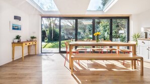 A modern kitchen with a large island, a dining area with a wooden bench and chairs, and a view of the garden through large sliding doors. - Trevint - Modern beach house in Falmouth, private parking (Falmouth)