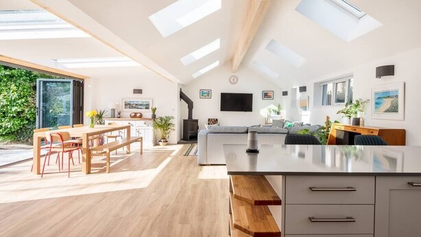A spacious living room with a large wooden dining table, a sofa, and a TV mounted on the wall. There are skylights and a large window providing natural light. - Trevint - Modern beach house in Falmouth, private parking (Falmouth)