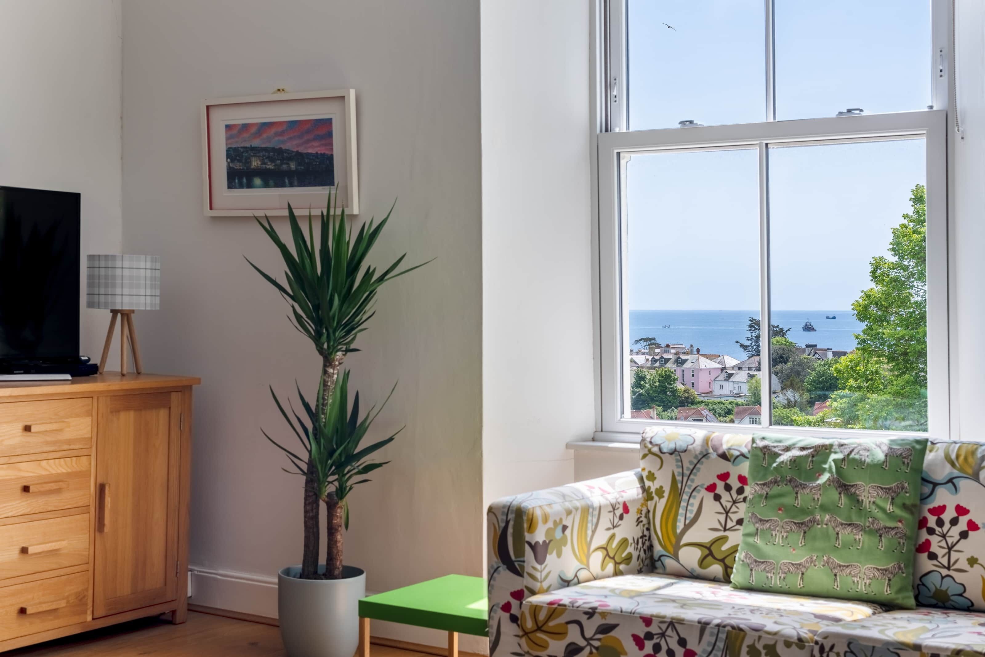 Een kamer met een houten kast, een potplant, een groene bank, een bank met bloemenkussens en een groot raam met uitzicht op zee en gebouwen.