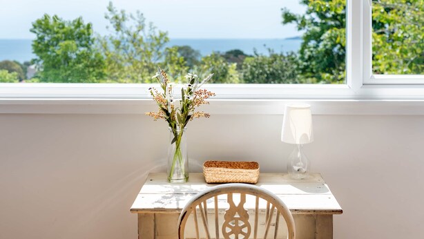 Una silla de madera orientada hacia una mesa pequeña con un florero, una vela y una cesta tejida, cerca de una ventana con vistas a la vegetación y al mar.