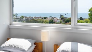 Un dormitorio con dos camas, una mesita de noche con lámpara y vistas al mar y a las casas a través de una ventana.