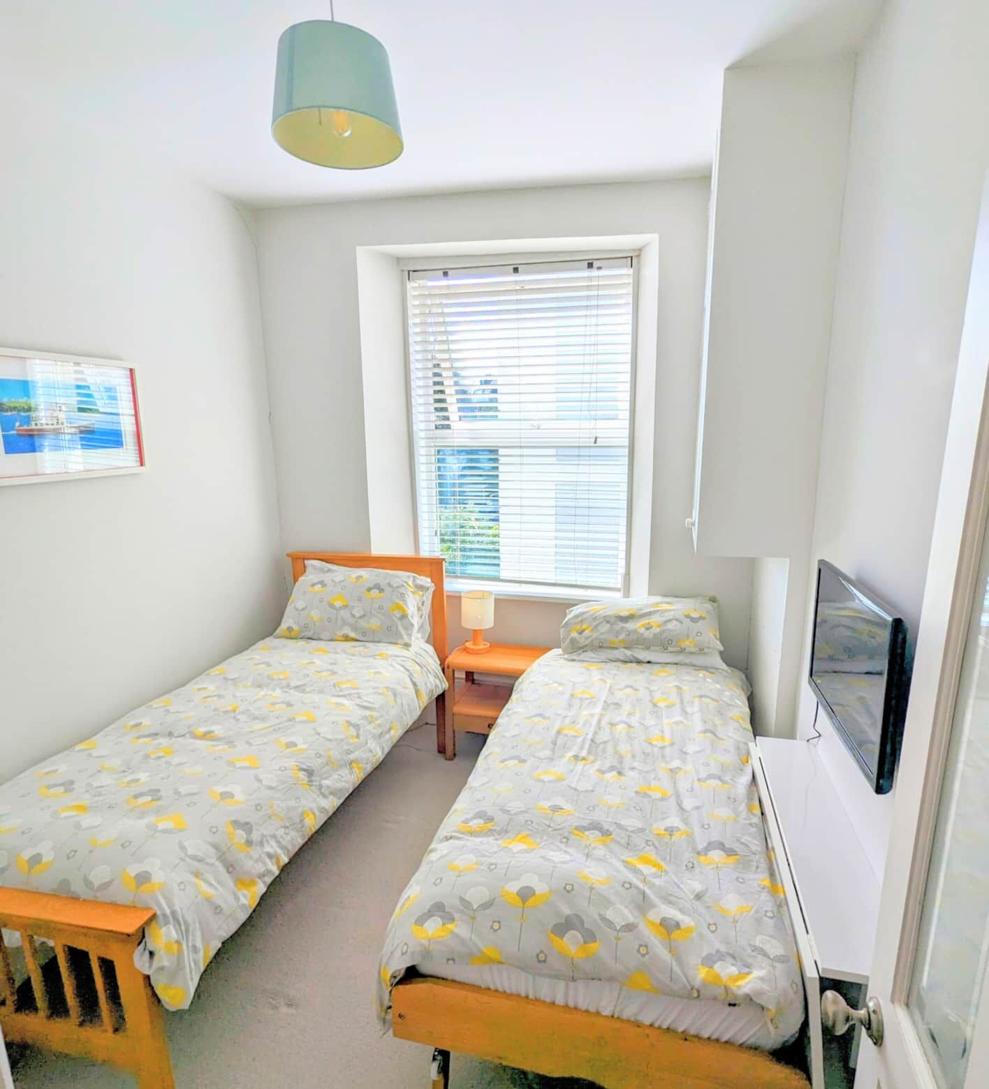 A small, well-lit bedroom with two single beds, a window with blinds, a desk, and a TV mounted on the wall.