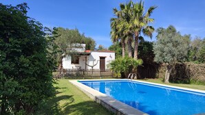 A swimming pool with a wooden deck, surrounded by lush greenery and a white building with a red roof. - Andalucía; Cádiz; Vejer de la Frontera;  cottage  Finca la Hijuela, casa Encina (Vejer de la Frontera)