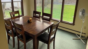 A dining area with a wooden table and chairs, large windows with blinds, and a standing fan. - Quiet single-level fully furnished house (Bangor)