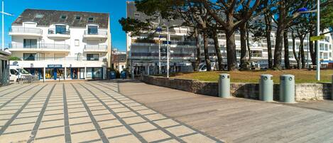 Uma passarela pavimentada com padrão quadriculado, que leva a um edifício com várias varandas e uma fachada comercial.