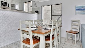 A dining area with a wooden table set for four, surrounded by white chairs with wooden seats. A microwave and oven are visible in the background.