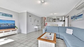 A modern living room with a sofa, coffee table, and TV. There is a kitchen area with cabinets and appliances.