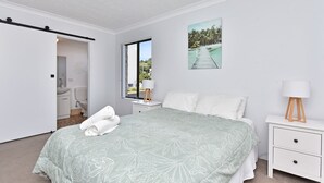 Un dormitorio con cama, mesita de noche, ventana con vista a palmeras y puerta que da a un baño.