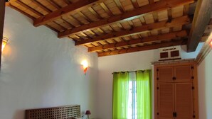 A bedroom with a wooden ceiling, a bed with patterned bedding, a nightstand, a lamp, a wardrobe, and green curtains. - Finca la Hijuela, Casa Azahar Andalucía; Cádiz; Vejer de la Frontera; (Vejer de la Frontera)
