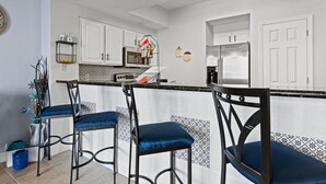 A kitchen with white cabinets, a stainless steel refrigerator, and a bar area with blue cushioned chairs. - Gorgeous Ocean and Bayside View (Miramar Beach)