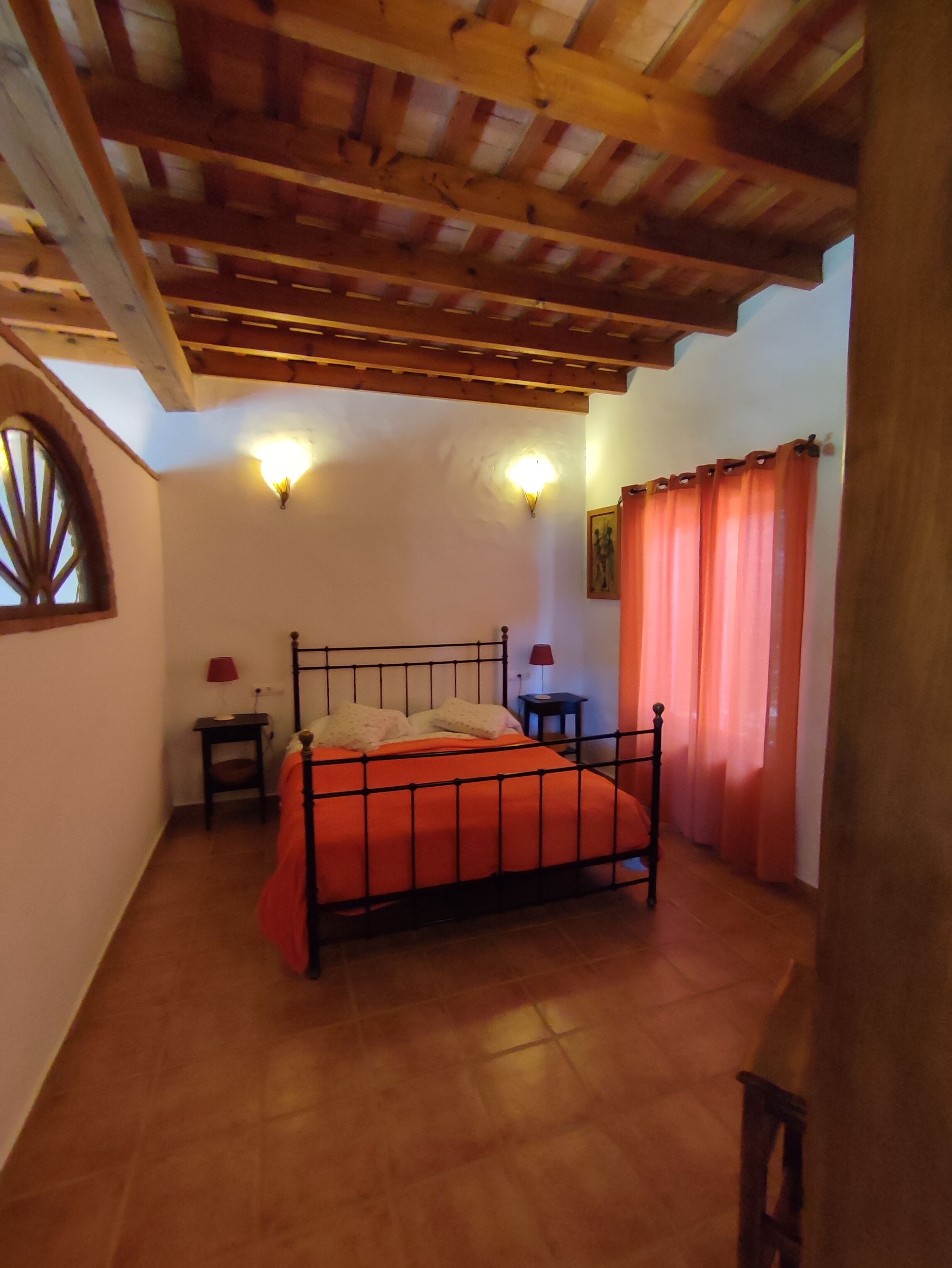 A bedroom with a wooden ceiling, a bed with an orange bedspread, two bedside tables with lamps, and a window with curtains.