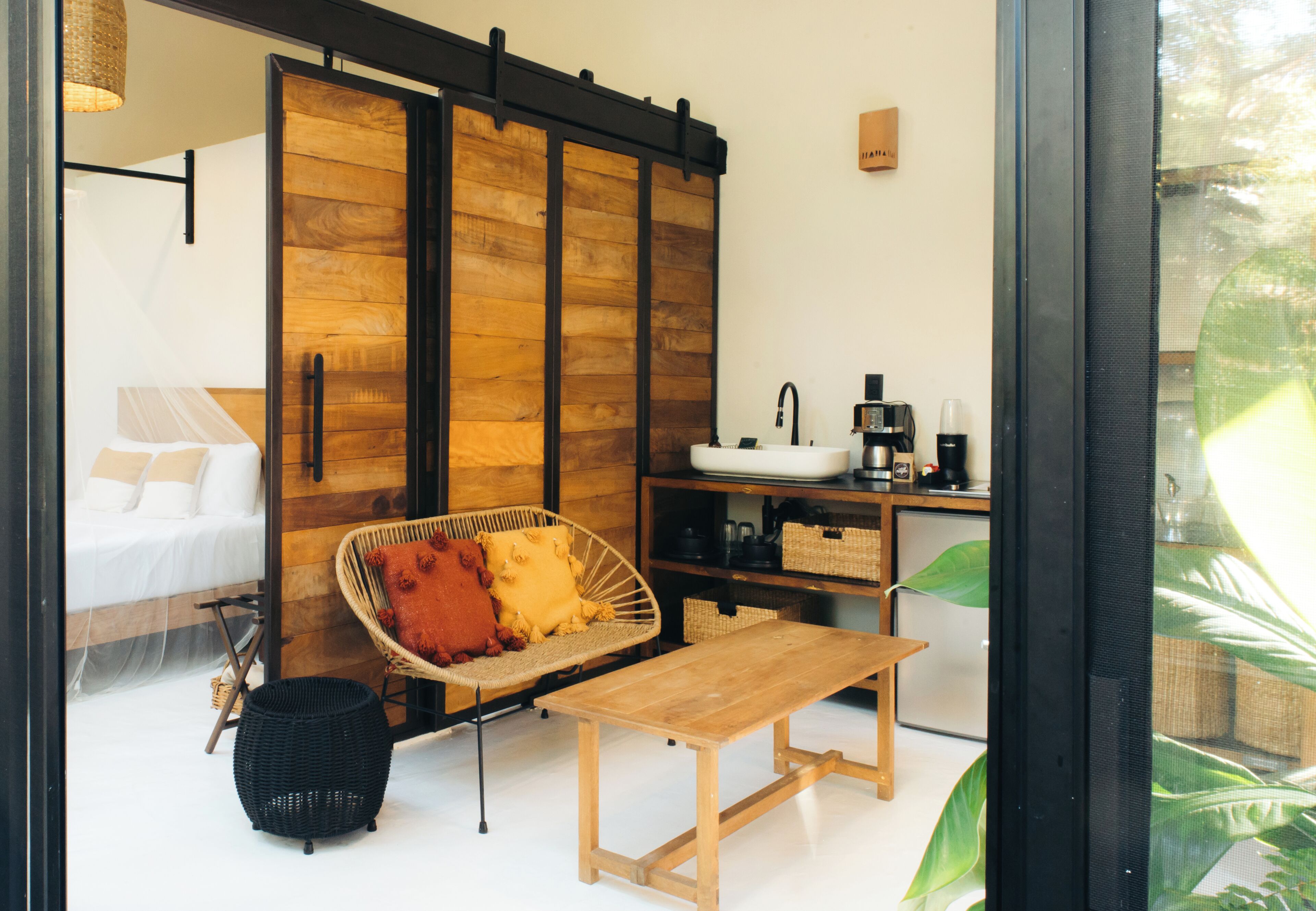 A cozy room with a bed, a wicker chair, a wooden table, a kitchenette, and a sliding door.